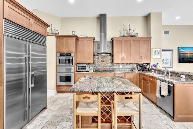 kitchen with sink, wall chimney exhaust hood, stainless steel appliances, light stone counters, and a kitchen island