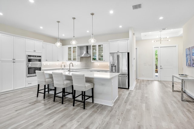 kitchen featuring an island with sink, decorative light fixtures, light hardwood / wood-style flooring, wall chimney range hood, and appliances with stainless steel finishes