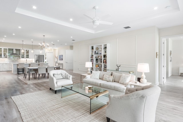 living room featuring ceiling fan with notable chandelier, a tray ceiling, and light wood-type flooring