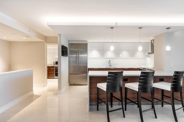 kitchen with tasteful backsplash, decorative light fixtures, wall chimney exhaust hood, built in fridge, and a kitchen breakfast bar