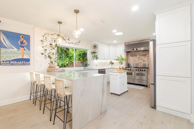 kitchen featuring decorative light fixtures, wall chimney range hood, a kitchen island, appliances with stainless steel finishes, and white cabinets