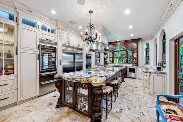 kitchen featuring light stone counters, ornamental molding, decorative light fixtures, stainless steel built in refrigerator, and a kitchen island with sink