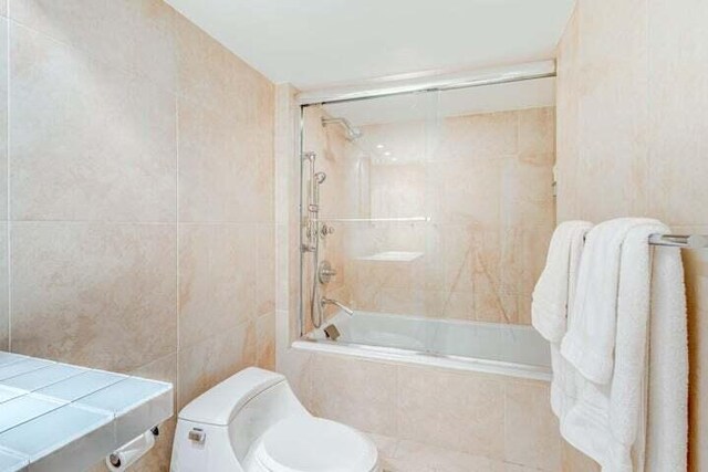 bathroom with toilet, tile walls, and bath / shower combo with glass door