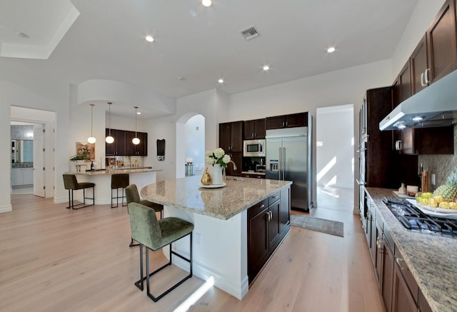 kitchen featuring built in appliances, hanging light fixtures, dark brown cabinetry, light stone counters, and a kitchen island