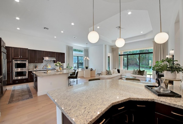 kitchen featuring stainless steel double oven, a large island with sink, light stone counters, and decorative light fixtures