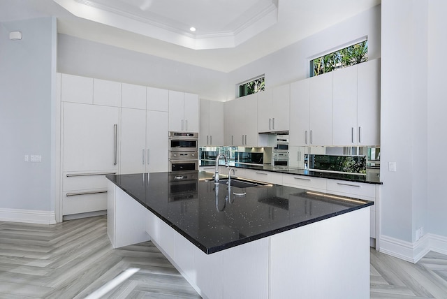 kitchen with crown molding, a tray ceiling, a center island with sink, sink, and white cabinets