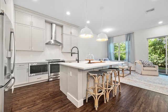 kitchen with a kitchen island with sink, wall chimney range hood, appliances with stainless steel finishes, decorative light fixtures, and dark hardwood / wood-style flooring