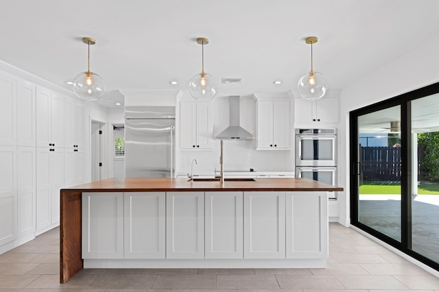 kitchen featuring an island with sink, appliances with stainless steel finishes, wooden counters, and wall chimney range hood