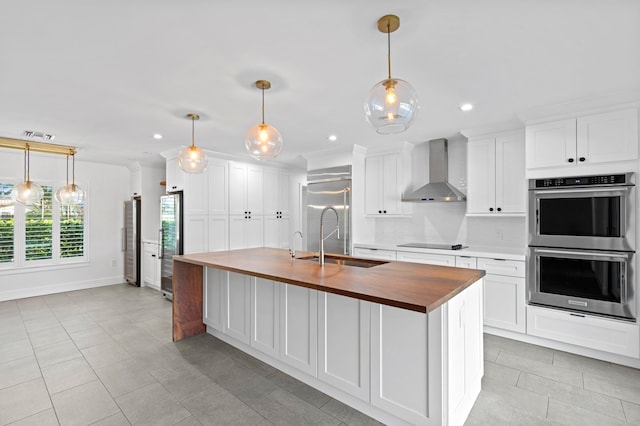 kitchen featuring appliances with stainless steel finishes, an island with sink, white cabinets, butcher block countertops, and wall chimney range hood