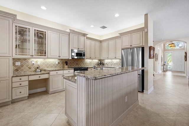 kitchen with light tile patterned flooring, a center island with sink, stainless steel appliances, light stone countertops, and decorative backsplash