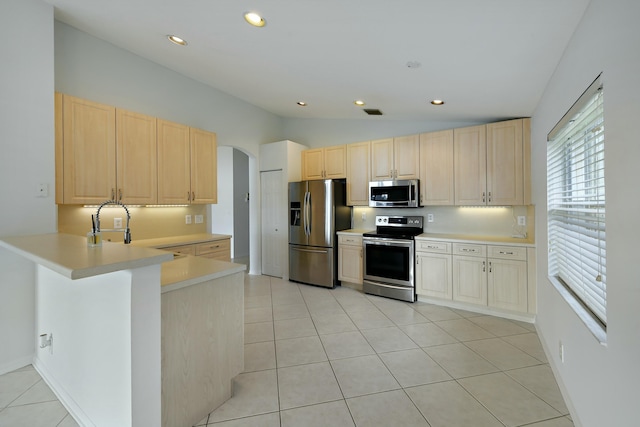 kitchen with kitchen peninsula, appliances with stainless steel finishes, light brown cabinetry, vaulted ceiling, and light tile patterned floors