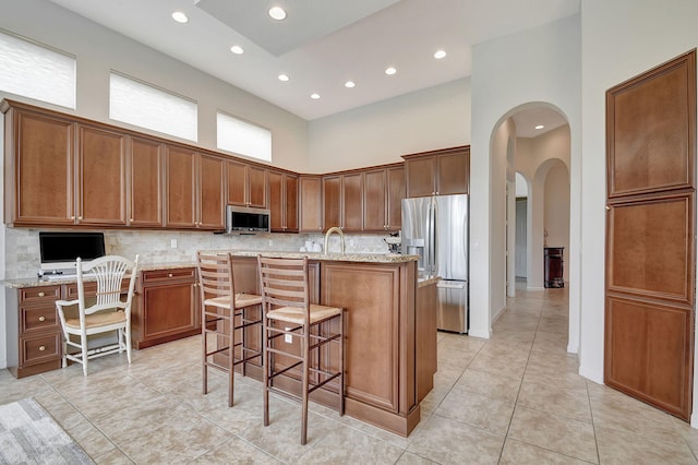 kitchen with an island with sink, stainless steel appliances, light stone countertops, a towering ceiling, and a kitchen bar