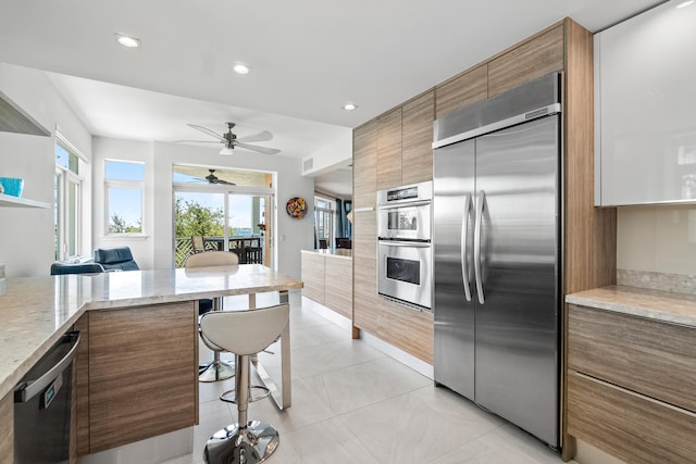 kitchen featuring appliances with stainless steel finishes, light tile patterned flooring, light stone countertops, a breakfast bar area, and ceiling fan