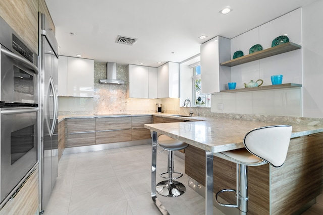 kitchen with white cabinets, stainless steel double oven, a kitchen bar, kitchen peninsula, and wall chimney exhaust hood