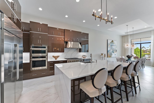 kitchen with appliances with stainless steel finishes, dark brown cabinets, decorative light fixtures, a kitchen island with sink, and sink