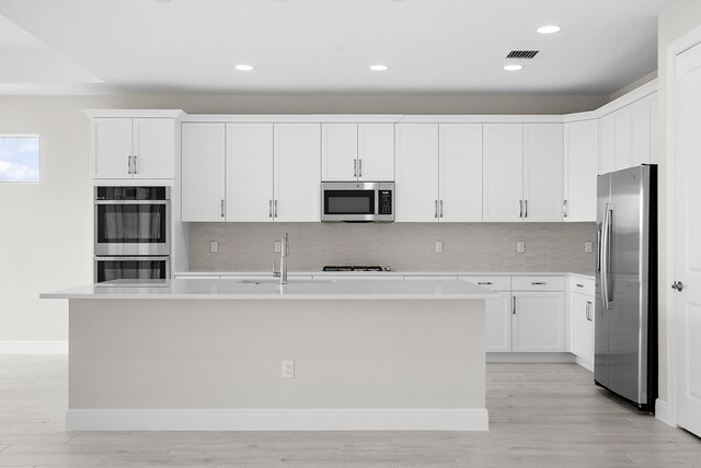 kitchen with appliances with stainless steel finishes, white cabinets, backsplash, light hardwood / wood-style flooring, and a kitchen island with sink