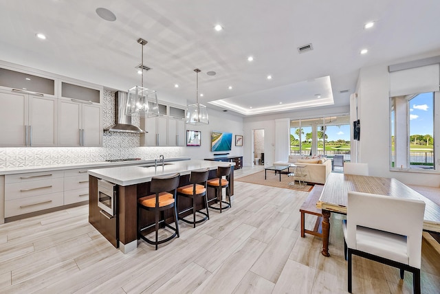 kitchen with pendant lighting, a tray ceiling, an island with sink, wall chimney range hood, and appliances with stainless steel finishes