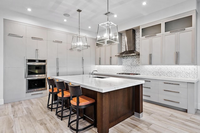 kitchen with pendant lighting, stainless steel appliances, a kitchen island with sink, sink, and wall chimney range hood