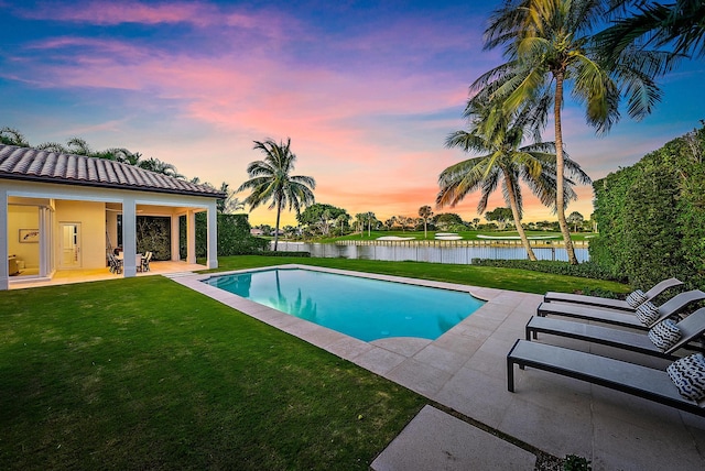 pool at dusk featuring a lawn, a water view, and a patio area