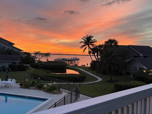 pool at dusk with a water view, a patio area, and a lawn