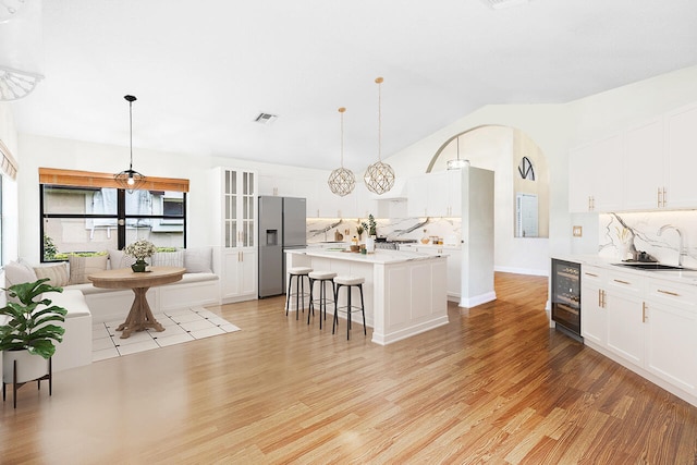 kitchen featuring wine cooler, light wood-type flooring, stainless steel refrigerator with ice dispenser, hanging light fixtures, and an island with sink