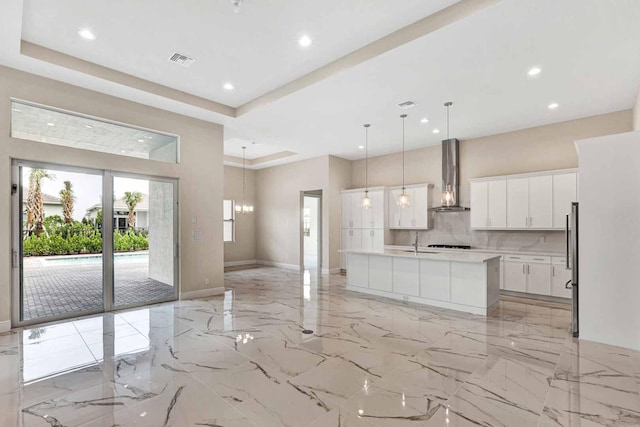 kitchen with an island with sink, backsplash, white cabinetry, wall chimney exhaust hood, and decorative light fixtures