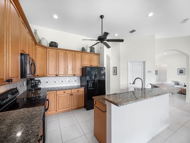 kitchen featuring light tile patterned flooring, black appliances, ceiling fan, dark stone counters, and a kitchen island with sink