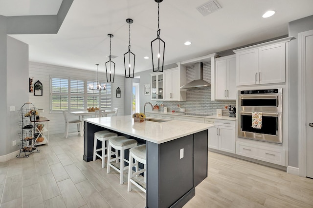 kitchen with white cabinets, wall chimney exhaust hood, stainless steel double oven, sink, and decorative light fixtures