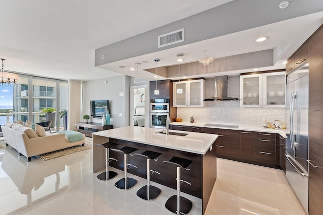 kitchen featuring a kitchen island with sink, hanging light fixtures, wall chimney range hood, decorative backsplash, and stainless steel appliances