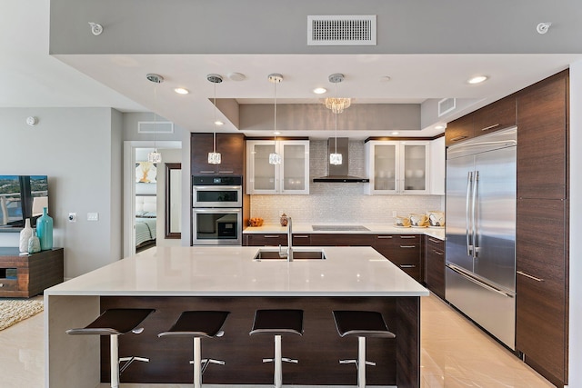 kitchen with an island with sink, sink, wall chimney range hood, appliances with stainless steel finishes, and a breakfast bar