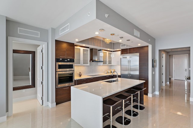 kitchen with appliances with stainless steel finishes, dark brown cabinetry, decorative light fixtures, a center island with sink, and wall chimney range hood