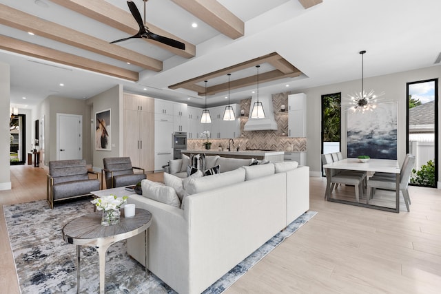 living room with sink, ceiling fan with notable chandelier, a raised ceiling, beamed ceiling, and light hardwood / wood-style flooring