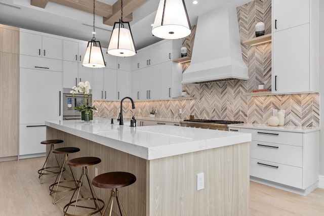 kitchen featuring light wood-type flooring, an island with sink, white cabinetry, pendant lighting, and custom exhaust hood