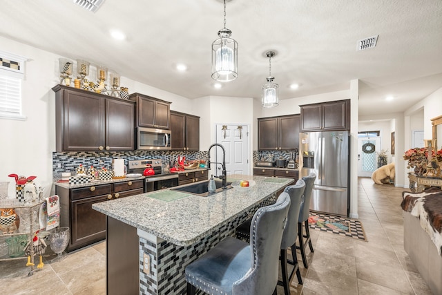 kitchen featuring tasteful backsplash, a kitchen island with sink, stainless steel appliances, dark brown cabinetry, and decorative light fixtures