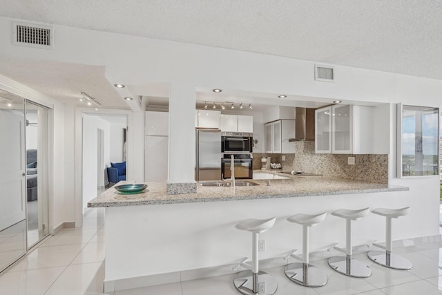 kitchen with a textured ceiling, white cabinets, kitchen peninsula, wall chimney range hood, and stainless steel appliances