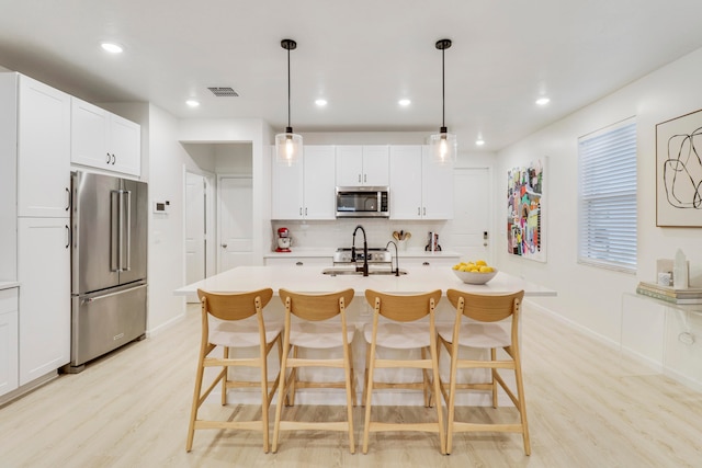 kitchen featuring a kitchen island with sink, appliances with stainless steel finishes, light wood-type flooring, and hanging light fixtures