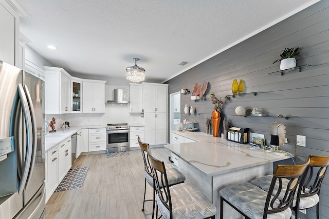 kitchen featuring a breakfast bar, white cabinets, wall chimney exhaust hood, stainless steel appliances, and light wood-type flooring