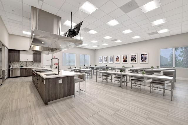 kitchen featuring a kitchen breakfast bar, dark brown cabinets, backsplash, a center island with sink, and a drop ceiling