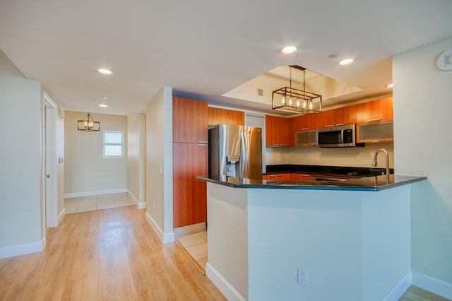 kitchen with a raised ceiling, dark countertops, appliances with stainless steel finishes, brown cabinets, and a peninsula