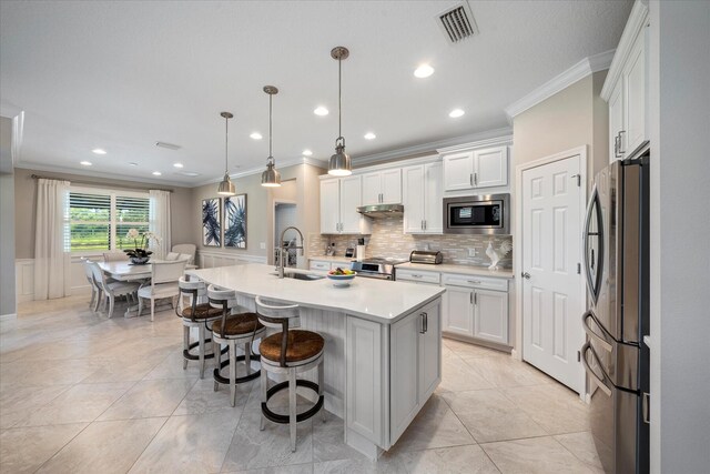 kitchen with a kitchen island with sink, white cabinets, hanging light fixtures, stainless steel appliances, and ornamental molding