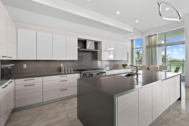 kitchen with tasteful backsplash, sink, white cabinets, a large island with sink, and wall chimney exhaust hood