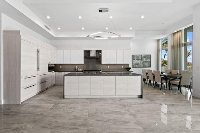 kitchen featuring white cabinetry, an island with sink, backsplash, stainless steel oven, and wall chimney exhaust hood