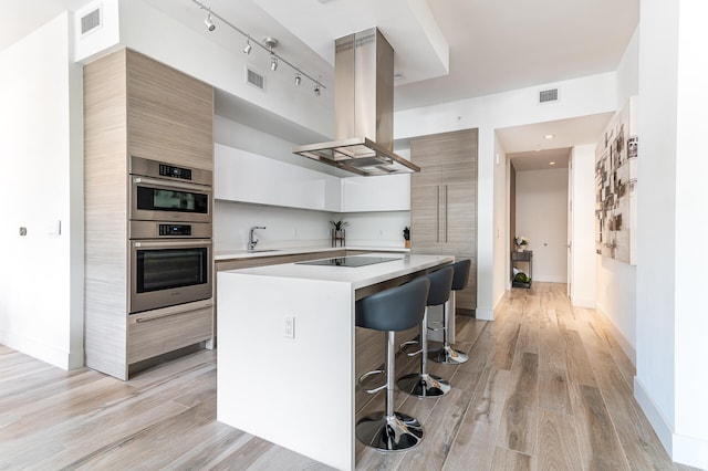 kitchen featuring light wood-type flooring, island exhaust hood, a kitchen bar, black electric cooktop, and double oven