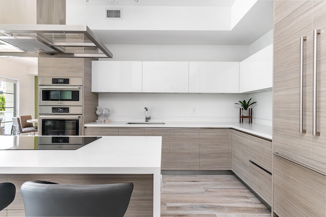 kitchen featuring a breakfast bar area, sink, white cabinetry, island range hood, and double oven