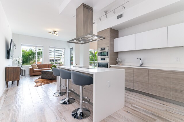 kitchen featuring sink, island exhaust hood, light hardwood / wood-style flooring, white cabinetry, and a breakfast bar