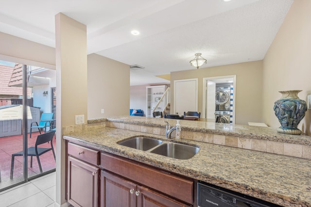 kitchen with black dishwasher, light stone countertops, light tile patterned floors, stacked washer and dryer, and sink