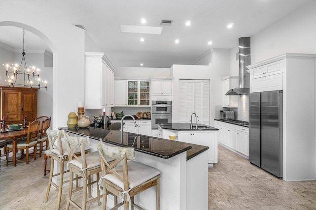 kitchen featuring wall chimney exhaust hood, a kitchen island with sink, appliances with stainless steel finishes, an inviting chandelier, and a kitchen bar