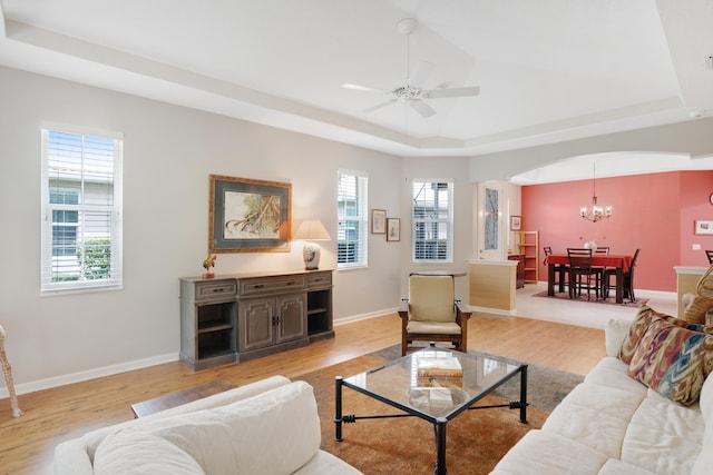 living room with ceiling fan with notable chandelier, a tray ceiling, light hardwood / wood-style floors, and plenty of natural light