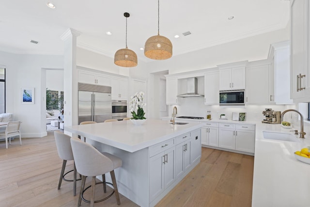 kitchen with built in appliances, wall chimney exhaust hood, white cabinets, and sink