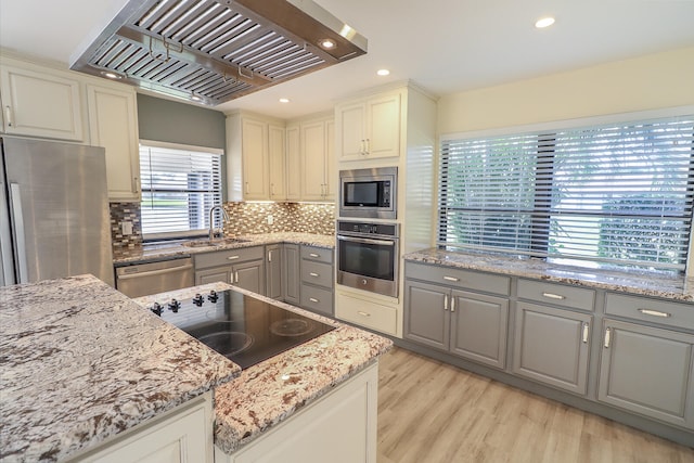 kitchen with gray cabinetry, exhaust hood, light stone countertops, appliances with stainless steel finishes, and light hardwood / wood-style floors